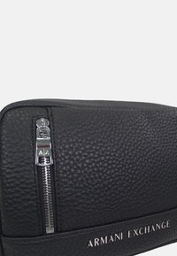 Fekete bőr táska texturált felülettel, ezüst cipzáras hardverrel és dombornyomott "Armani Exchange" logóval az elülső részen. Téglalap alakú.
