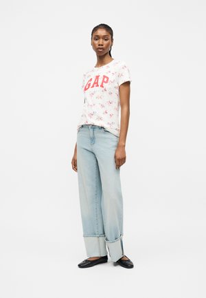 Femme portant un t-shirt blanc à motifs floraux avec le logo "GAP", un jean large bleu clair retroussé aux chevilles et des ballerines noires, debout devant un fond blanc.