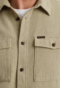 Chemise à motif chevron beige avec deux poches poitrine, boutons marron et étiquette PME Legend. Conçue avec un col classique et un tissu texturé.