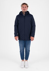 Marineblaue Jacke mit Kapuze, Reißverschluss vorne und langen Ärmeln, kombiniert mit hellblauen Jeans und weißen Turnschuhen. Glatte Stofftextur.