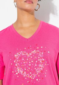 Camiseta de cuello en V rosa con pequeños corazones dispersos en varias tonalidades de rosa dentro de una forma de corazón. Material de algodón suave.
