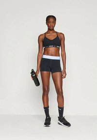 Soutien-gorge de sport noir avec logo blanc, short noir assorti avec une ceinture de taille bleu clair, chaussettes noires et sneakers noirs, tenant une bouteille d'eau.