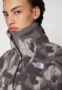 Pullover in pile grigio con motivi astratti neri. Presenta un colletto alto, una zip frontale a metà e un logo ricamato bianco sul petto.