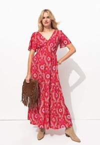 Robe maxi rouge à fleurs avec des manches courtes volantées, taille cintrée et décolleté en V. Assortie à un sac à franges marron et des bottines beiges.