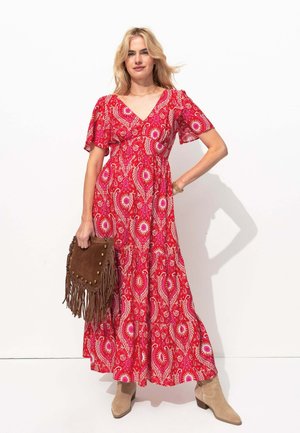 Robe maxi rouge à fleurs avec des manches courtes volantées, taille cintrée et décolleté en V. Assortie à un sac à franges marron et des bottines beiges.