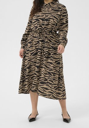 Femme portant une robe boutonnée à manches longues aux rayures tigre noires et beiges, nouée à la taille, associée à des chaussures noires pointues.