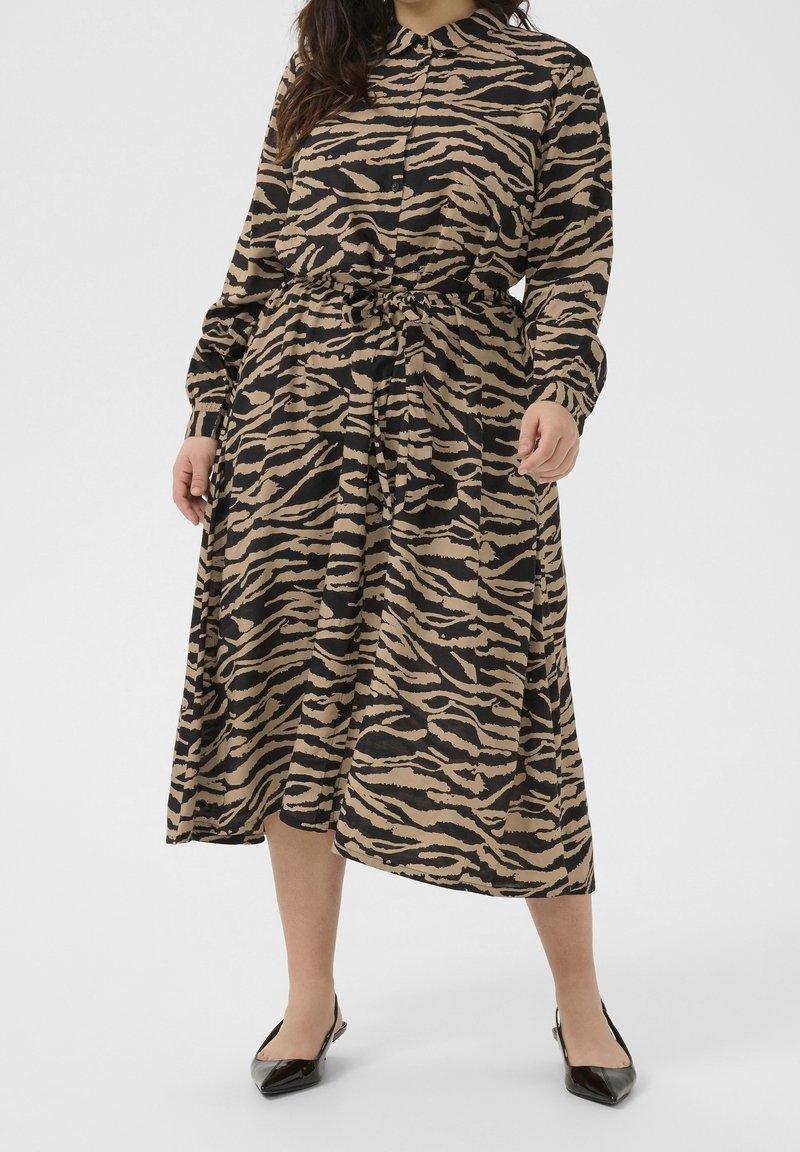 Femme portant une robe boutonnée à manches longues aux rayures tigre noires et beiges, nouée à la taille, associée à des chaussures noires pointues.