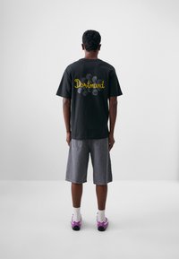 Svart t-skjorte i bomull med "Dortmund" i gul kursiv bakpå, parret med grå denimshorts og lilla sneakers. Enkel bakgrunn.