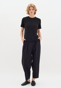 Melnā īsām piedurknēm t-krekls ar smalku tekstūru, saskaņots ar brīvām melnām biksēm un melniem slip-on apaviem, demonstrēts uz baltā fona.