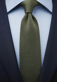 Stickad grön slips med ett strukturerat mönster, bärs över en ljusblå skjorta, under en mörkblå blazer.