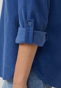 Chemise en velours côtelé bleu avec des manches retroussées, dotée d'une fermeture à boutons et d'un tissu texturé. L'ourlet est légèrement courbé.