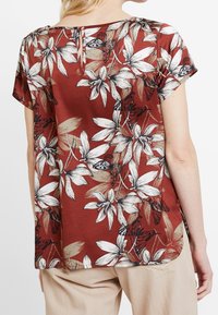 Kortærmet, rustfarvet top med hvid blomster- og sommerfuglemønster. Stoffet er glat med en subtil glans og har en nøglehuldetalje på ryggen.