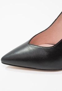 Chaussure à talon haut en cuir noir à bout pointu avec une texture lisse et une doublure intérieure rose pâle.