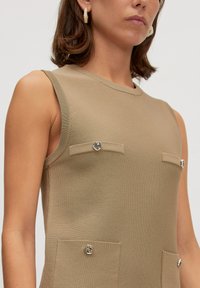 Beige ärmelloses Rippenoberteil mit zwei vorderen Taschen, die jeweils mit einem dekorativen silbernen Knopf verziert sind. Glatte Textur, figurnahes Design.