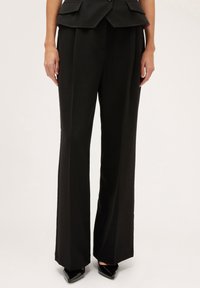 Pantalones anchos de corte entallado en negro, con una textura suave, que presentan una cintura alta y pliegues prensados para mayor detalle. Combinados con zapatos negros de punta.