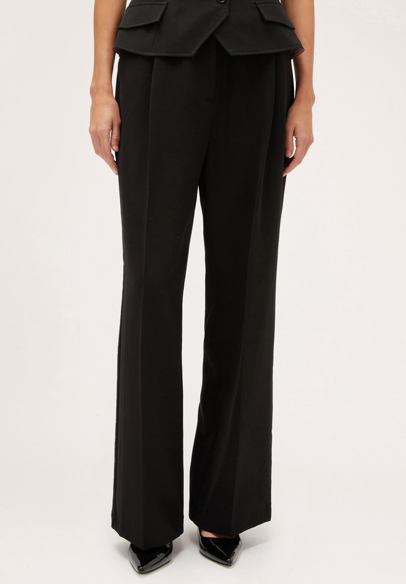 Pantalones anchos de corte entallado en negro, con una textura suave, que presentan una cintura alta y pliegues prensados para mayor detalle. Combinados con zapatos negros de punta.