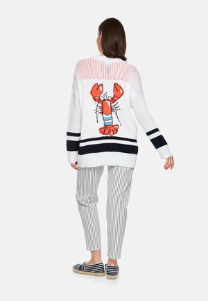 Woman wearing white knit sweater with large red lobster design on back, striped white pants, and navy striped espadrilles, standing facing away.