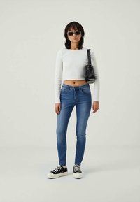 Camisola cropped de manga longa branca, jeans ajustados azuis, sapatilhas pretas e brancas, mala de ombro preta e óculos de sol azuis sobre um fundo neutro.