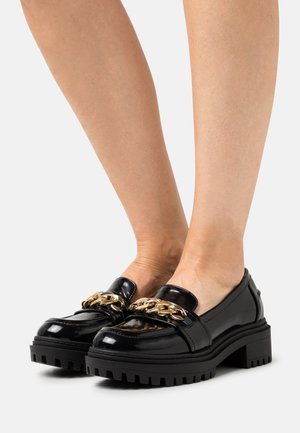 Mocassins en cuir noir brillant avec des semelles épaisses et un détail en chaîne dorée portés pieds nus sur fond blanc uni.