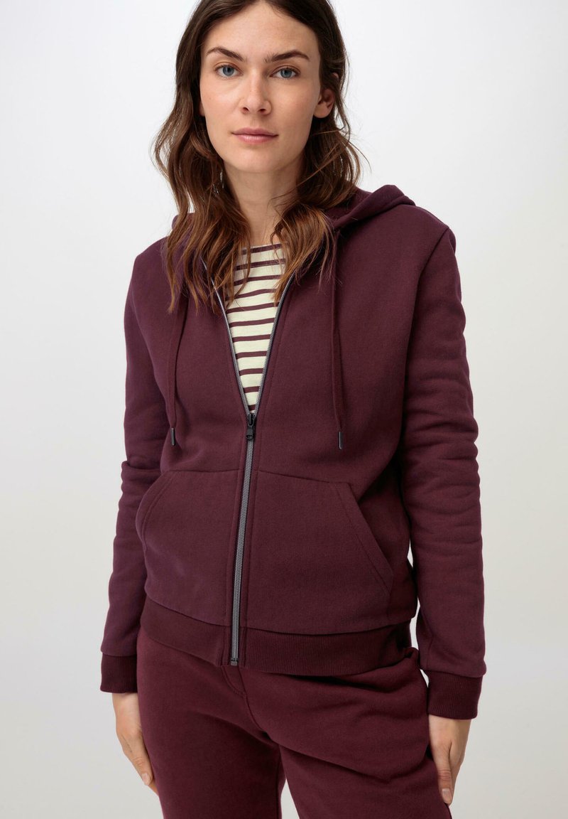 Bordeauxfarbener Reißverschluss-Hoodie mit Kapuze, Fronttaschen und gerippten Bündchen, getragen über einem gestreiften navyblauen und weißen Shirt. Weicher, strukturierter Stoff.