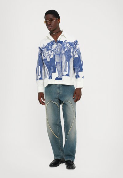 Uomo in jeans di denim blu con grandi toppe a forma di cuore sfrangiate e una camicia bianca con una stampa di una folla blu, in piedi su uno sfondo bianco.