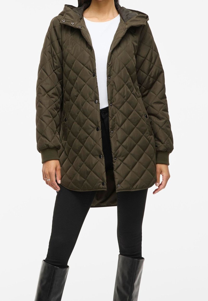Femme portant une veste matelassée vert olive avec des boutons-pression, un pantalon noir, des bottes noires et une chemise blanche en dessous.