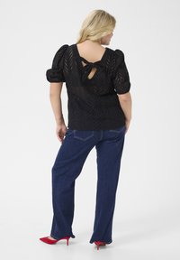 Black lace blouse with puff sleeves and back tie detail, paired with dark blue jeans and red high heels. White background.