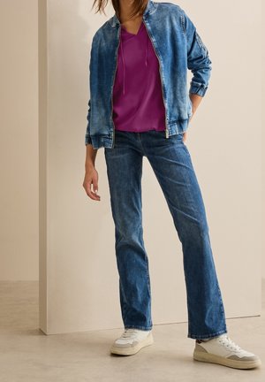 Personne portant une veste en jean bleue, un top violet, un jean bleu droit et des baskets blanches, debout devant un fond beige uni.