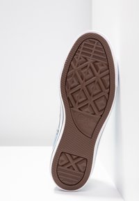 Semelle d'une basket classique Converse All Star montrant une bande de roulement en caoutchouc marron avec des motifs géométriques et un logo, reposant sur un fond blanc.