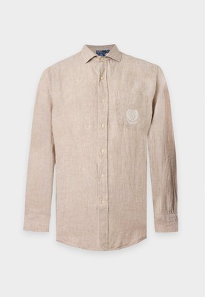 Jeune homme portant une chemise beige boutonnée, un pantalon gris foncé et des baskets blanches, debout avec une main dans la poche.