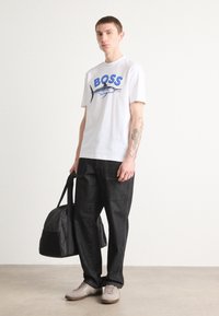 Camiseta blanca de manga corta con texto azul de "BOSS" y gráfico de marlín, combinada con pantalones de mezclilla oscuros y zapatillas beige, sosteniendo un bolso negro.