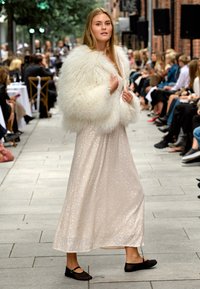 Lang, glitrende beige kjole med paljetter, kombinert med en hvit, fluffy pelsfrakk. Svarte, flate sko fullfører antrekket. Motebane-setting.