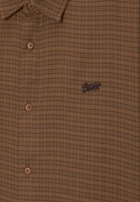 Camisa de cuadros marrón hecha de tela suave, con cuello con solapa, cinco botones y un logotipo bordado en azul oscuro.