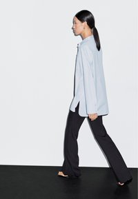Light blue button-up shirt with a relaxed fit and side slits, paired with black flared pants and flat sandals, against a minimalist background.