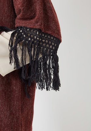 Persona que lleva un chal de textura burdeos con flecos negros sobre un bolso beige claro, contra un fondo liso.