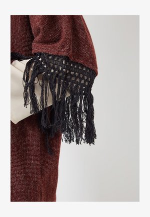 Persona que lleva un chal de textura burdeos con flecos negros sobre un bolso beige claro, contra un fondo liso.