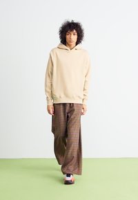 Hanorac bej cu glugă, cu manșete ribbate, combinat cu pantaloni largi din fibră de carouri, având nuanțe de verde, maro și roșu. Modelul poartă pantofi sport.