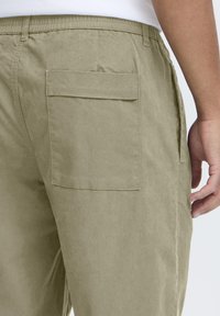 Pantalon en coton vert olive clair avec une poche latérale, une taille élastique et une texture lisse avec des coutures visibles sur les côtés.