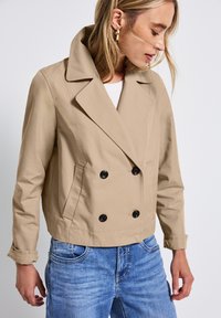Beige dvouřadový kabát s velkými černými knoflíky, klopovým límcem a bočními kapsami. Tkanina vypadá hladce a je mírně strukturovaná.