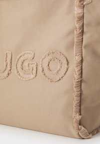 Bež torba od tkanine sa strukturnom vezom koja čita "HUGO," sa raspucanim ivicama i glatkom površinom. Kvadratnog oblika sa ojačanim uglovima.