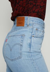 Jeans de ganga azul claro com cintura alta, apresentando um remendo de marca em couro castanho, bolsos traseiros com um padrão de costura distinto e uma pequena etiqueta vermelha.