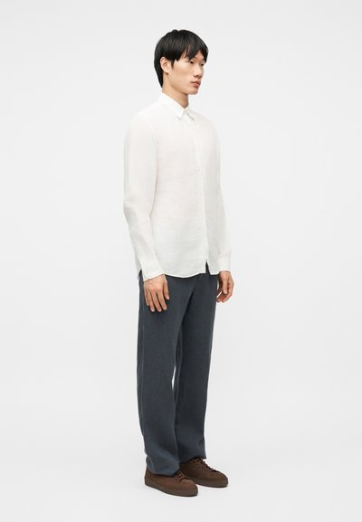 Άνδρας όρθιος, φορώντας λευκό πουκάμισο με μακριά μανίκια και κουμπιά, σκούρο γκρι παντελόνι και καφέ παπούτσια, μπροστά σε απλό ανοιχτό φόντο.