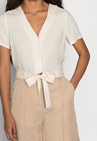 Mujer con una blusa beige de manga corta abotonada atada en un lazo a la cintura, combinada con pantalones de tiro alto color marrón claro.
