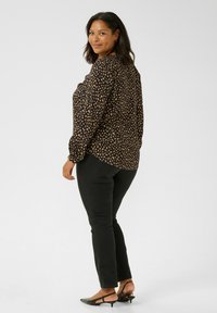 Black patterned blouse with long sleeves, featuring a subtle design in shades of tan. Paired with fitted black pants and low-heeled shoes.