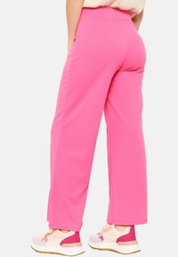 Pantalon large rose en tissu doux et extensible. Présente une taille haute et une texture lisse. Porté avec des chaussures de sport multicolores.