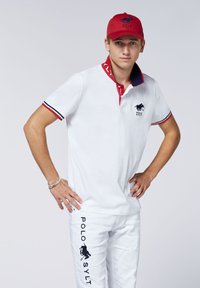 Weißes Poloshirt mit roten und marineblauen Akzenten an Kragen und Ärmeln. Weiße Hose mit dem Text und Logo "POLO SYLT". Rote Kappe mit Logo auf der Vorderseite.