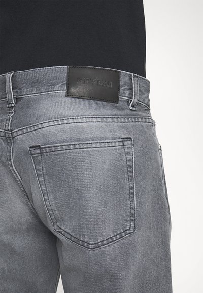 Jeans de ganga cinzentos com um rótulo de marca em pele preta na cintura, com dois bolsos traseiros e uma leve textura desgastada.