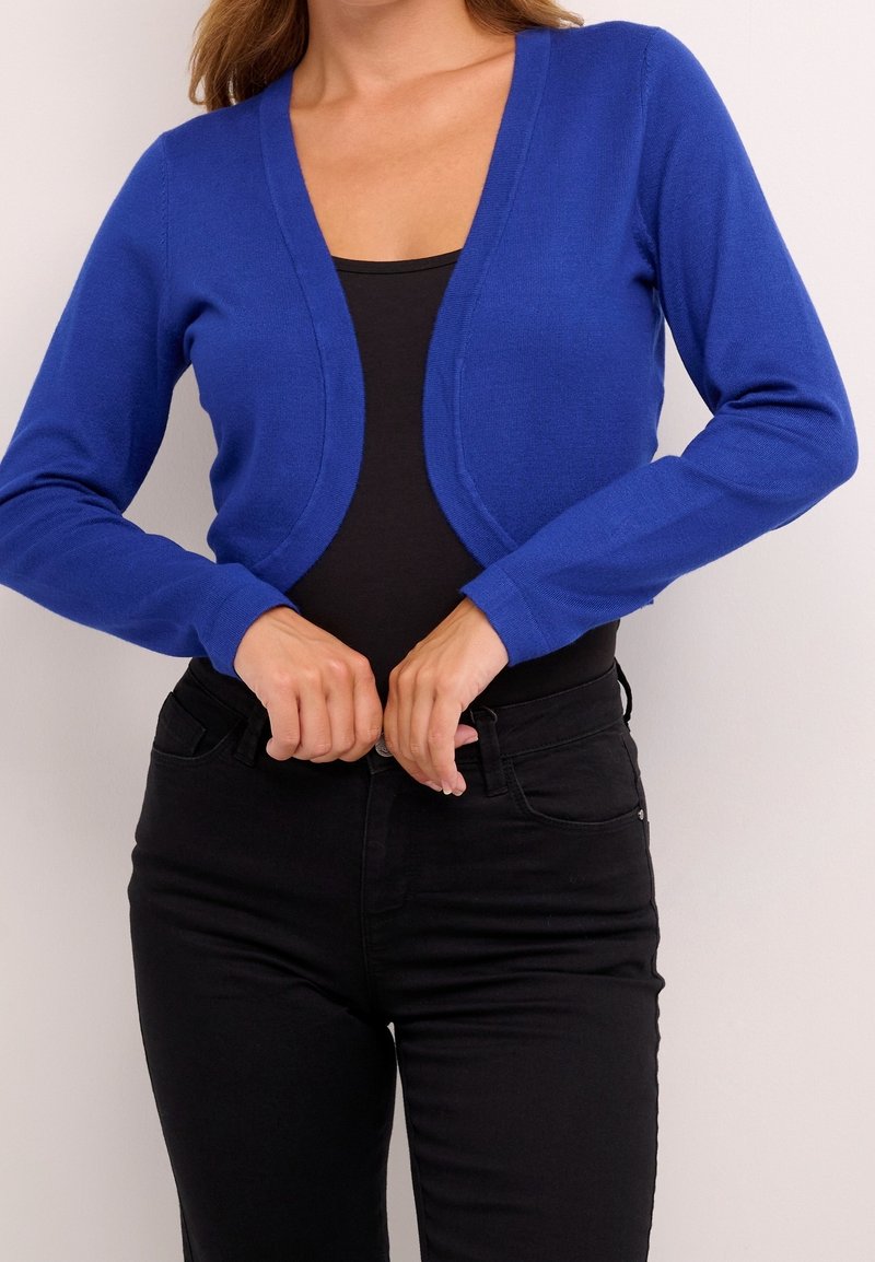 Cardigan bleu en maille avec un col en V profond et des manches longues, tissu texturé, porté sur un haut noir et assorti à un jean noir.