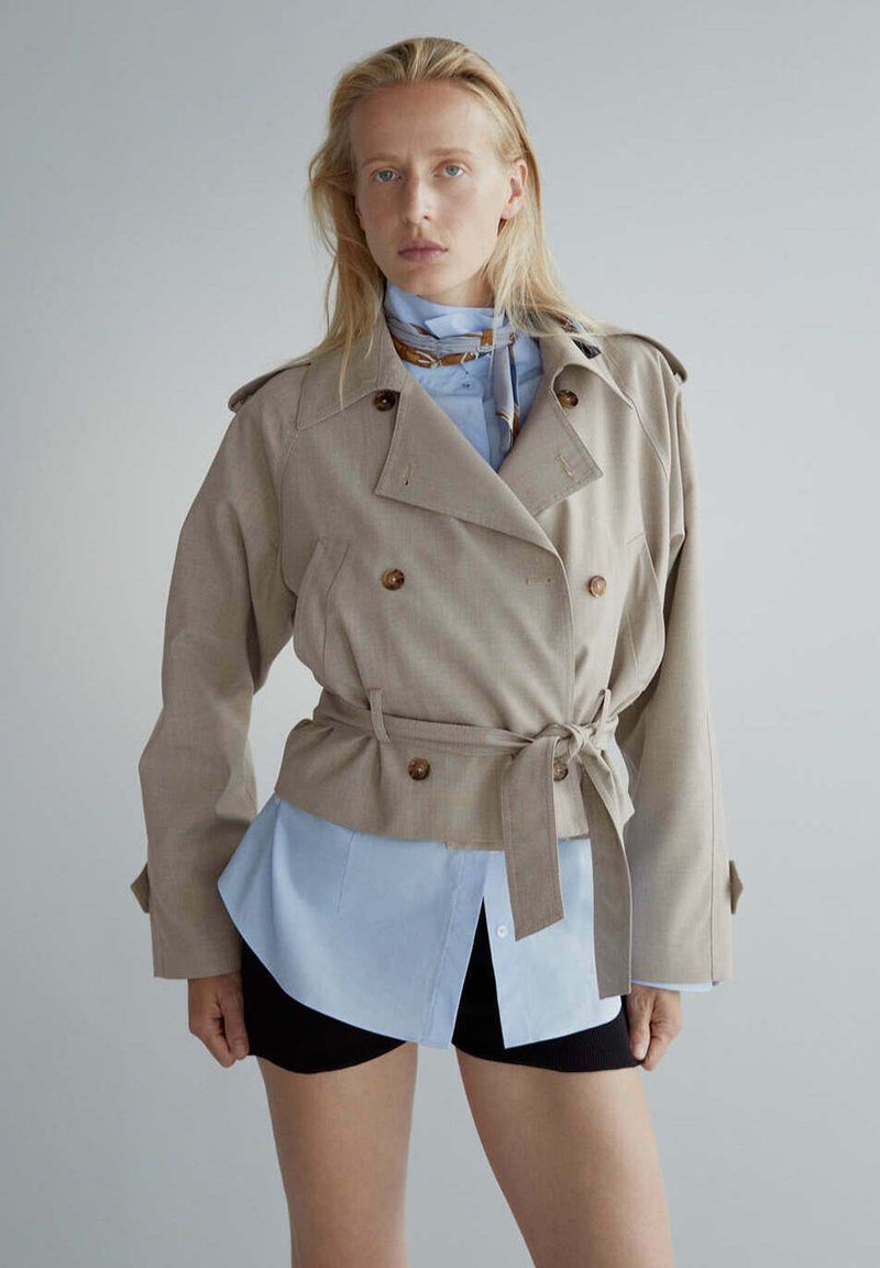 Femme portant une veste beige ceinturée sur une chemise bleu clair et un short noir, debout devant un fond gris uni.
