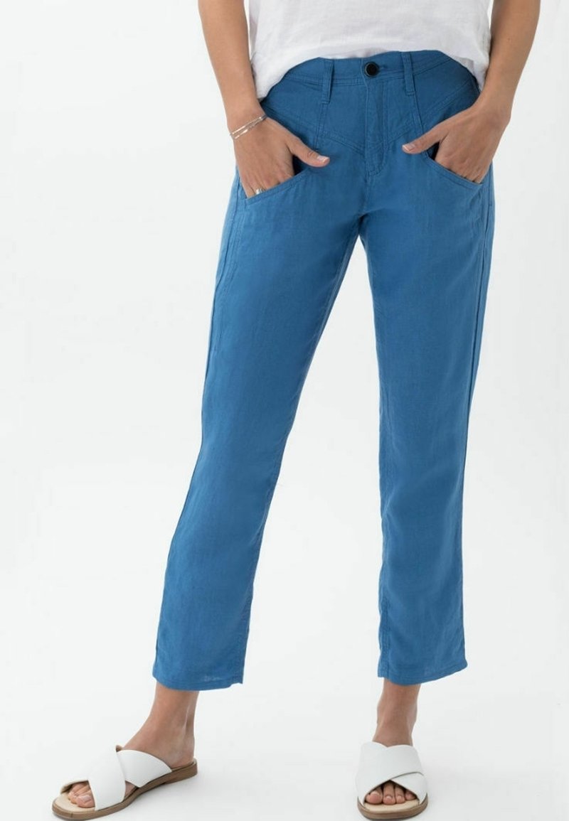 Personne portant un pantalon bleu à jambes droites, les mains dans les poches avant et des sandales blanches sans bride sur fond blanc uni.